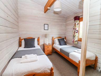 A bedroom with two single beds a nightstand with a lamp a window with curtains and wooden walls at Snowy Owl Lodge in Rhayader