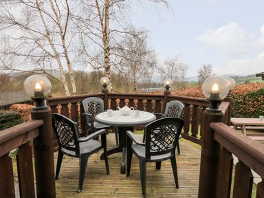 A wooden deck with a round table and four plastic chairs surrounded by wooden railing with globe lights at Snowy Owl Lodge in Rhayader