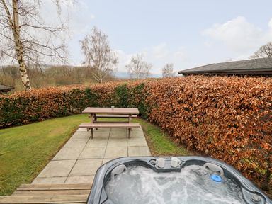 An outdoor area with a bubbling hot tub in foreground a wooden picnic table on a paved patio and hedges surrounding the lawn at Snowy Owl Lodge in Rhayader