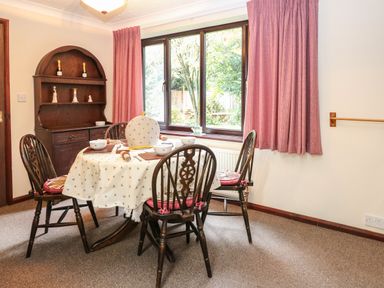 A dining area with a wooden table and chairs set for a meal near a window with pink curtains at Walnut Tree Cottage in Catfield