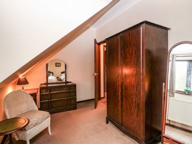 A bedroom corner with a wooden wardrobe a mirror dresser a chair a side table and a lamp at Walnut Tree Cottage in Catfield