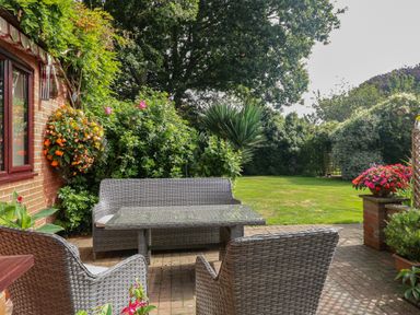 A garden patio with wicker chairs and a table surrounded by green bushes and flowers at Walnut Tree Cottage in Catfield