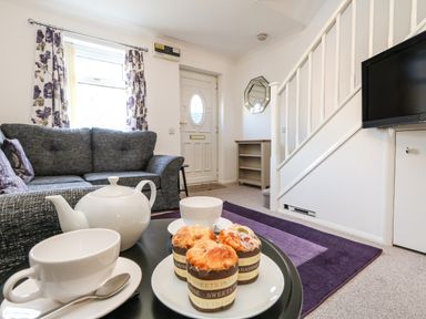 A living room with a gray sofa white teapot cups muffins a purple rug a wall-mounted tv and white stairs at Kingfisher Cottage in Weymouth