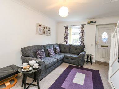 A living room with a gray sectional sofa and purple cushions a black side table with tea set and cupcakes a black footrest a purple rug a window with floral curtains framed hearts wall art and a white front door at Kingfisher Cottage in Weymouth