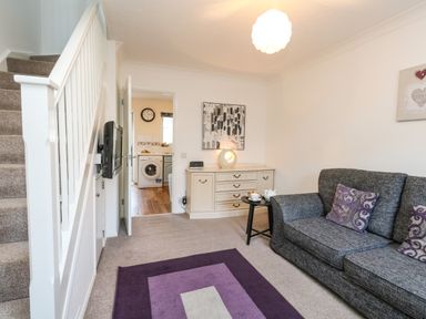 A living room with a gray sofa with purple pillows a purple rug a white staircase and a sideboard at Kingfisher Cottage in Weymouth