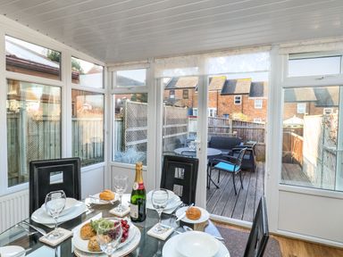 A dining area with a glass table set with plates glasses and a bottle overlooking a wooden deck with outdoor furniture at Kingfisher Cottage in Weymouth