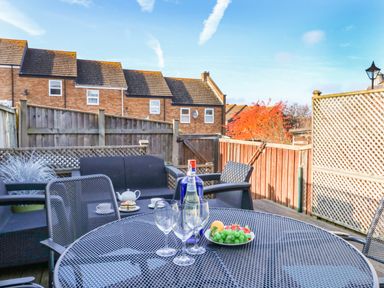 An outdoor patio with a metal table and chairs bottles glasses and fruit with houses and a wooden fence in the background at Kingfisher Cottage in Weymouth