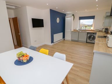 A kitchen with a white table and colorful chairs a fruit bowl on the table a washing machine and cabinets along one wall Berth in Afonwen near Criccieth