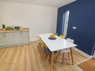 A dining area with a white table surrounded by mixed color chairs and a fruit basket on top next to kitchen cabinets at Berth in Afonwen near Criccieth