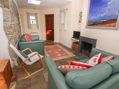 A living room with stone wall a wooden door two green sofas a rocking chair a fireplace and paintings at Rospletha Bungalow in Treen