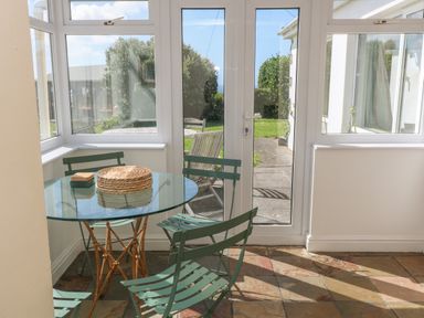 A dining area with a round glass table and green metal chairs near glass doors at Rospletha Bungalow in Treen