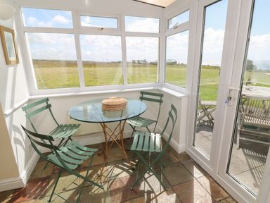 A sunroom with a glass round table and four green metal chairs overlooking a grassy field at Rospletha Bungalow in Treen