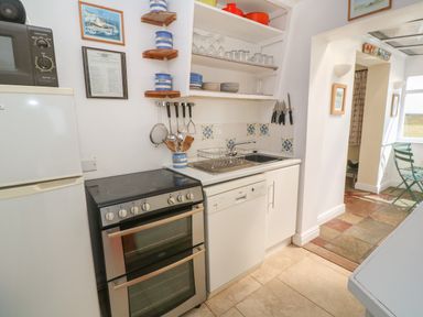 A kitchen with a refrigerator, stove, dishwasher, sink, open shelves with dishes and glassware, and kitchen utensils at Rospletha Bungalow in Treen