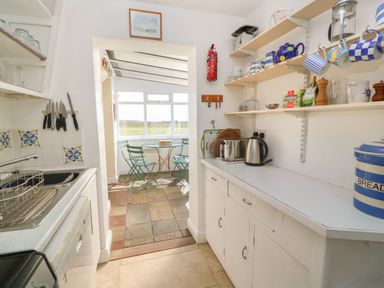 A kitchen with shelves holding mugs and kitchen items and a small dining area with a round table and chairs at Rospletha Bungalow in Treen