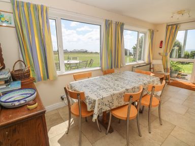 A dining area with a table covered by a patterned tablecloth and six chairs with windows showing a garden view at Rospletha Bungalow in Treen