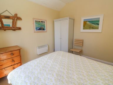 A bedroom with a bed covered in a floral bedspread a wooden chest of drawers a white wardrobe a wooden chair and framed paintings on the wall at Rospletha Bungalow in Treen