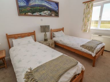 A bedroom with two single beds a nightstand with a lamp a window with curtains and a landscape painting on the wall at Rospletha Bungalow in Treen