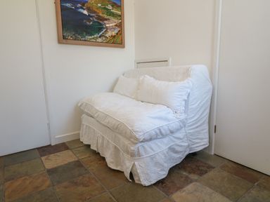 A small white armchair with cushions on a tiled floor against white walls with a painting above at Rospletha Bungalow in Treen