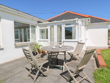 An outdoor patio with a round wooden table and five wooden chairs outside a white bungalow at Rospletha Bungalow in Treen
