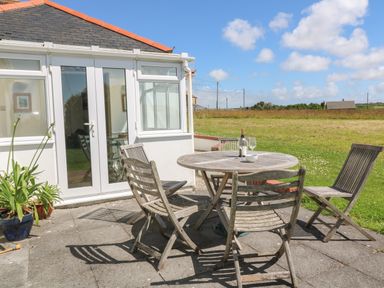 A patio with a round wooden table and four wooden chairs outside a white house with glass doors and windows at Rospletha Bungalow in Treen