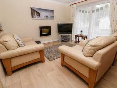 A living room with two beige sofas a wall-mounted fireplace a television on a stand a small wooden table and large window with curtains at 17 River Banks in Liskeard