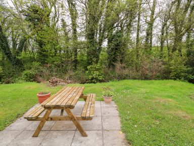 A wooden picnic table on a patio with potted plants and grass with trees in the background at 17 River Banks in Liskeard