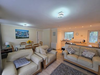 A living area with beige sofas and cushions a dining table with chairs and flowers and a kitchen with a window at 17 River Banks in Liskeard