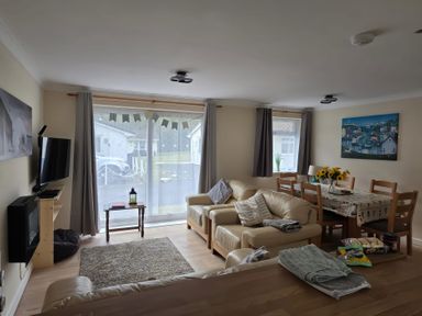 A living room with beige leather sofas a dining table with wooden chairs and sunflowers on it and a television on the wall at 17 River Banks in Liskeard