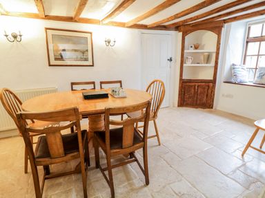 A dining room with a wooden table and six chairs a wall cabinet and a window with cushions at Teign Cottage in Shaldon