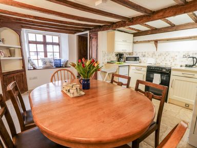 A kitchen with wooden ceiling beams a wooden dining table and chairs a window with cushions and kitchen appliances at Teign Cottage in Shaldon
