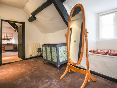 A room with a standing oval mirror wooden chest with floral panels a window with cushions and a doorway leading to another room at Teign Cottage Shaldon