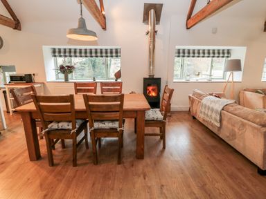 A dining area with wooden table and chairs, a stove with a chimney, windows with blinds, a sofa with a blanket, and a floor lamp at The Old Workshop in Middleton near Belford