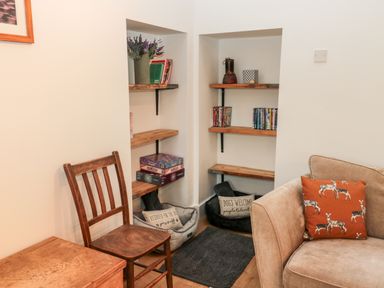A corner with wooden shelves holding books dvds and board games next to a wooden chair and a beige sofa with an orange pillow at The Old Workshop in Middleton near Belford