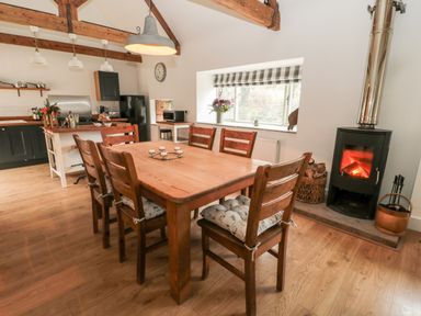 A dining area with a wooden table and chairs next to a wood burning stove and kitchen at The Old Workshop in Middleton near Belford