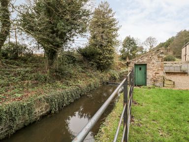 A small stone building with a green door beside a narrow waterway with metal railing and surrounding trees and grass at The Old Workshop in Middleton near Belford