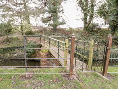 A metal gate and fence opening to a wooden footbridge over a stream with trees in the background at The Old Workshop in Middleton near Belford