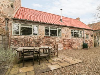 An outdoor stone patio with a wooden table and chairs next to a stone building with three windows and a red tiled roof at The Old Workshop in Middleton near Belford