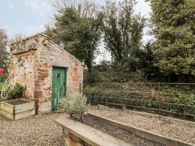 A small stone building with a green door beside a gravel path and garden beds at The Old Workshop in Middleton near Belford