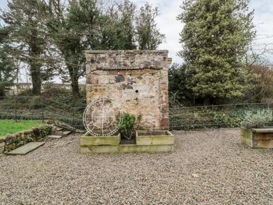 An outdoor area with a stone wall a metal clock frame wooden planters gravel ground and trees at The Old Workshop in Middleton near Belford
