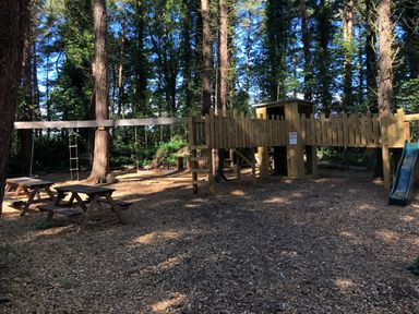 A playground with wooden climbing structures swings a slide and picnic tables in a wooded area at The Old Workshop in Middleton near Belford