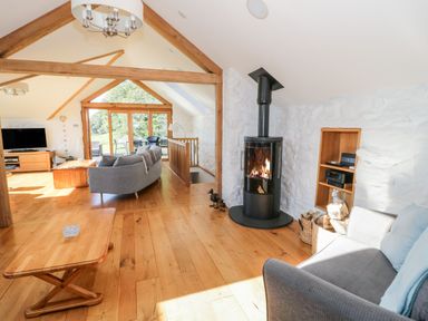 An open living room with wooden floors a gray sofa a wood stove and large windows at Tyn Llwyn Pencoed near Corwen