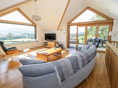 A living room with a curved sofa and armchair by large windows overlooking green fields at Tyn Llwyn in Pencoed near Corwen