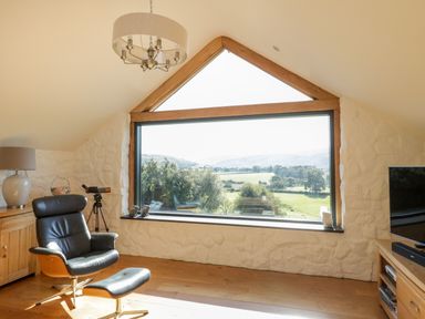 A living room with a large window overlooking countryside next to a black armchair and wooden furniture at Tyn Llwyn in Pencoed near Corwen