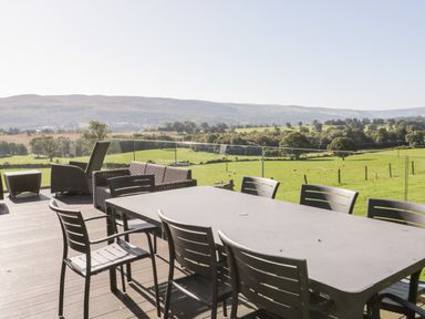 An outdoor dining table with chairs and lounge furniture on a wooden deck overlooking green fields and hills at Tyn Llwyn in Pencoed near Corwen