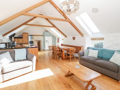 An open plan living and dining area with grey sofas wooden dining table and chairs a kitchen with wooden cabinets and a white fridge under wooden beams at Tyn Llwyn in Pencoed near Corwen