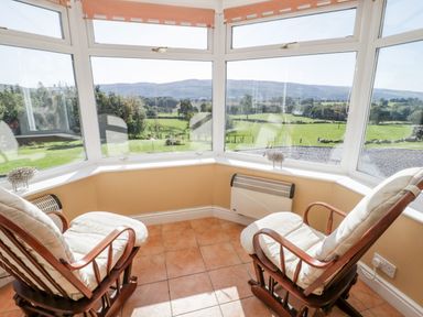 A sunroom with two cushioned wooden rocking chairs and large windows overlooking fields at Tyn Llwyn Pencoed near Corwen