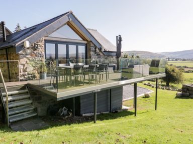 A stone house with a raised deck with glass railing and outdoor dining furniture at Tyn Llwyn in Pencoed near Corwen