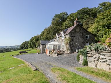 A stone house with a conservatory and a gravel driveway surrounded by green grass and trees at Tyn Llwyn in Pencoed near Corwen
