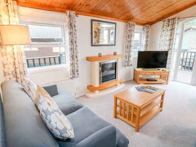 A living room with a grey sofa, wooden coffee table, television on wooden stand, fireplace, and windows with patterned curtains at Smugglers Shack in Polperro