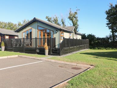 A single story cabin with a fenced deck next to a parking lot and grassy area
